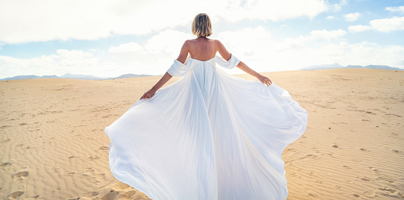 Back view of a blonde woman in fashionable, maxi white wedding dress posing on the desert, dancing in sunny light. Full length photo. Copy space.