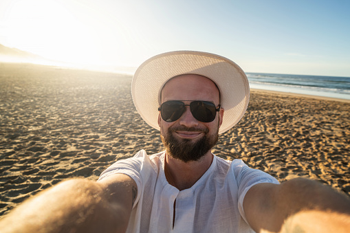 Happy, smiling man on vacation at the beach, taking selfie. Handsome guy in white summer hat and black sunglases.