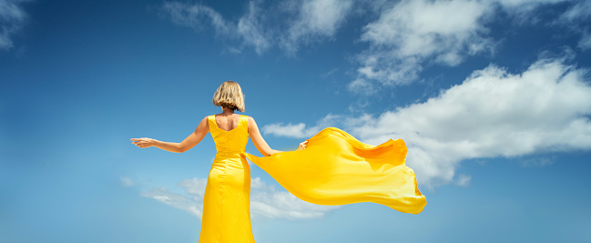 Fashionable blonde woman in yellow dress posing on the blue sky background, back view. A lot of copy space. Colors.