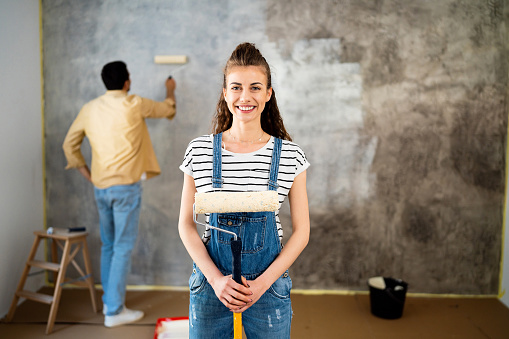 Portrait of a young Caucasian woman, with a paint roller in his hand, while her boyfriend paints the wall behind her in their new apartment