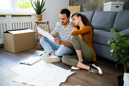 The young Caucasian couple, sitting on the floor eating fast food  and looking at the blueprint of the house, they moving in and renovating