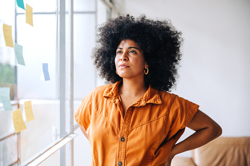 Pensive young businesswoman looking away while standing next to a glass wall in an office. Black female entrepreneur organising her creative ideas using adhesive notes.