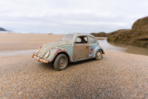 Perranporth, UK - 1st August, 2022:  A beaten and battered VW Beetle sits on a beach in Cornwall with the summer sunshine beating down on its faded paintwork. Scale model photography.