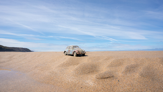 Perranporth, UK - 1st August, 2022:  A beaten and battered VW Beetle sits on a beach in Cornwall with the summer sunshine beating down on its faded paintwork. Scale model photography.
