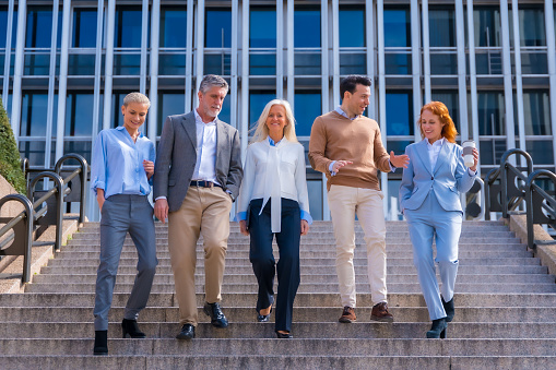 Cheerful group of coworkers outdoors in a corporate office area walking down some stairs going to work, middle-aged businesswoman and businessman