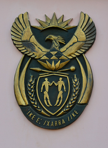 Bissau, Guinea-Bissau: National Coat of Arms of the Republic of South Africa, outside the embassy - It represents a shield with two people holding hands. A spear and a knobkierie (truncheon) cross above the shield, two ears of grain are on the left and right of the shield. This arrangement is surrounded by two tusks on each side, connected in a green band. Above the shield is a secretary bird with outstretched wings. In front of its chest is a protea flower, above his head is a rising sun. The motto is written in the extinct |Xam, member of the Khoisan languages, and translates literally to \