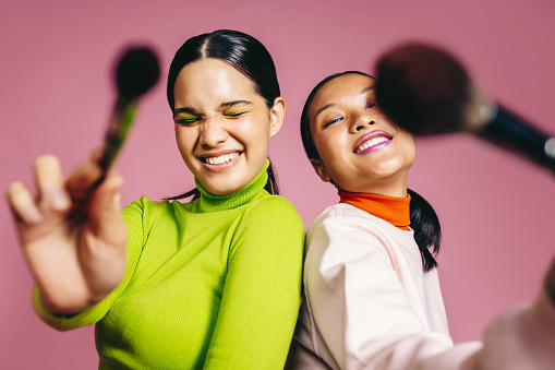 Females in their 20's smile happily while holding makeup brushes in a studio. Two glamorous young women females following trends by doing a social media challenge