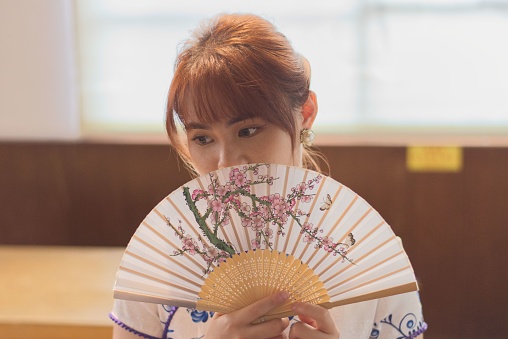 A young East Asian woman posing with an ornate oriental-style fan in front of her face.
