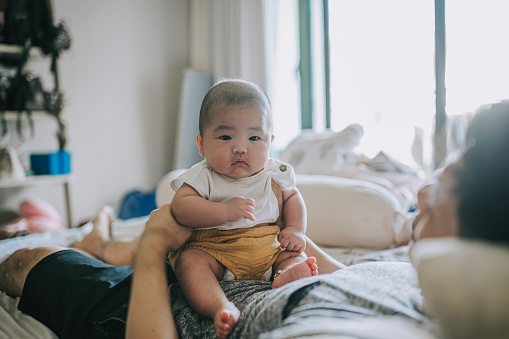 Asian Chinese baby boy sitting on father's abdomen on bed weekend morning