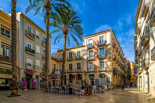 Alicante, Spain - JANUARY 17, 2020: Outdoor restaurant on small town square among palms and colorful buildings in old town of Alicante - city and historic Mediterranean port, famous tourist resort.