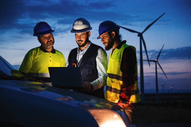 istock Team work of three renewable energy engineers with helmets and protection equipment, working and planing at night with laptop computer on top of the car with a wind power generators in the background. Renewable energy concept. 1485426289