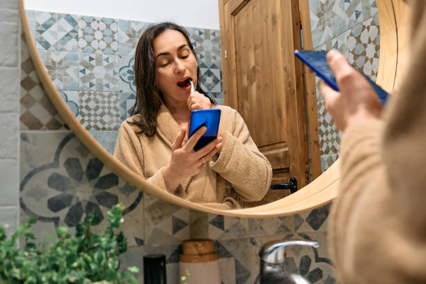 istock Woman brushing teeth with toothbrush and using smartphone for chatting and blogging in front of mirror in bathroom in the morning. Oral hygiene, healthy teeth and care. 1485440155