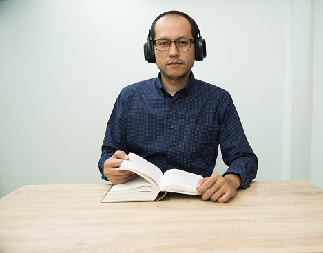 The man in deep blue shirt reading the book while on headset to listen music in library at university.