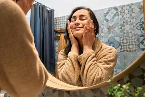 Middle-aged woman in bathrobe looking at mirror in bathroom and touching her face, applying hydrating anti-aging cosmetics on her skin and smiling.