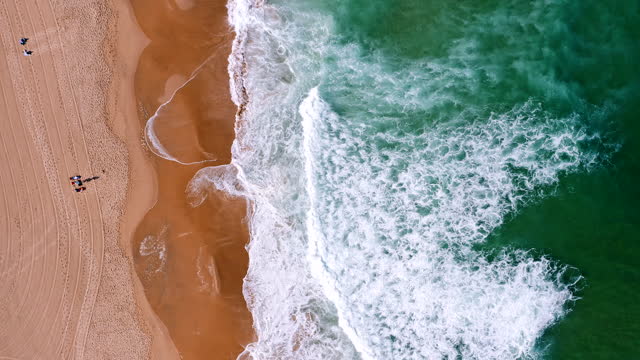 Top aerial view of waves break on tropical yellow sand beach. Bird's eye perspective of emerald green ocean surface and tourist relaxing on holiday