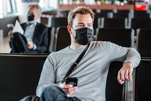 Waist up front shot with blurred background of a Caucasian mid-adult man seated waiting to board a flight at the airport gate wearing a black mask and looking away while holding his phone with the right hand.