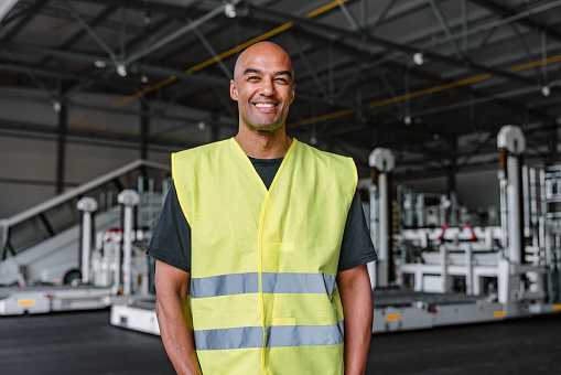 Medium shot with blurred background of a mid-adult Latin-American aeronautic engineer smiling and wearing a green and gray protective uniform as a part of his routine at the warehouse of the airport.