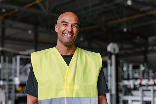 Medium shot with blurred background of a mid-adult Latin-American aeronautic engineer smiling and wearing a green and gray protective uniform as a part of his routine at the warehouse of the airport.