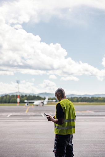 Three-quarter shot with blurred background of a mid-adult Latin-American aeronautic engineer checking a tablet and wearing a green and gray protective uniform as a part of his checking routine while he stands in front of a commercial airplane at the airport runway.