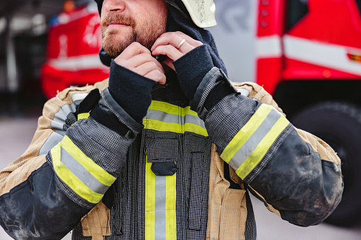 A Caucasian firefighter putting on his fire protection suit and helmet, standing next to a fire truck.