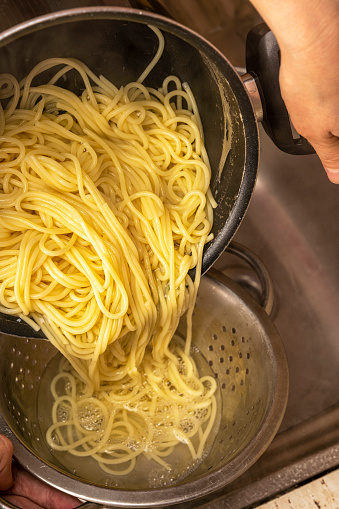 Colander with fresh cooked spaghetti. Woman pouring water from boiled spaghetti into colander. Spaghetti being poured into a colander. Pouring of water from boiled pasta over a kitchen sink.vertical photo