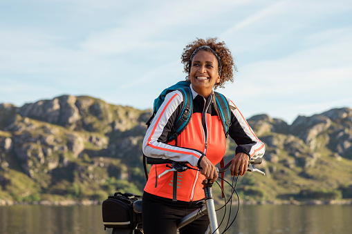 A woman on a bike is resting on her handlebars by the edge of a Scottish loch, she has on a rucksack and is smiling in the sunshine.