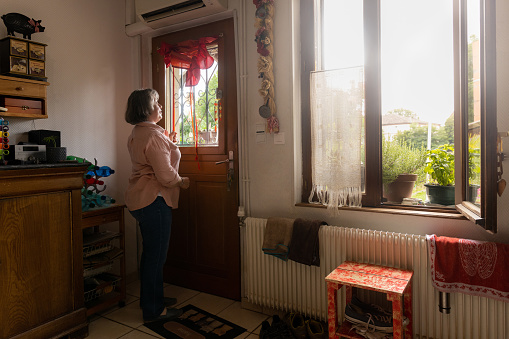 A mature woman who is blind, getting ready for the day ahead in her home in Castelferrus, France. She has just raised the blind up at her front door and is enjoying the  feeling of the warm sun on her face through the glass. The window in the room is open to let fresh air in.