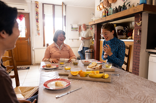 A woman sitting with her daughter and daughter's boyfriend at the kitchen table in their home kitchen in Castelferrus, France while the father of the family stands in the background. They are all laughing with each other and enjoying the morning together.