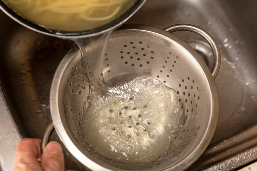 Colander with fresh cooked spaghetti. Woman pouring water from boiled spaghetti into colander. Spaghetti being poured into a colander. Pouring of water from boiled pasta over a kitchen sink.