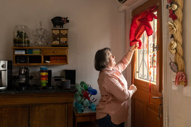 A mature woman who is blind, getting ready for the day ahead in her home in Castelferrus, France. She is raising the blind up at her front door and feeling the warmth of the morning sun on her face.