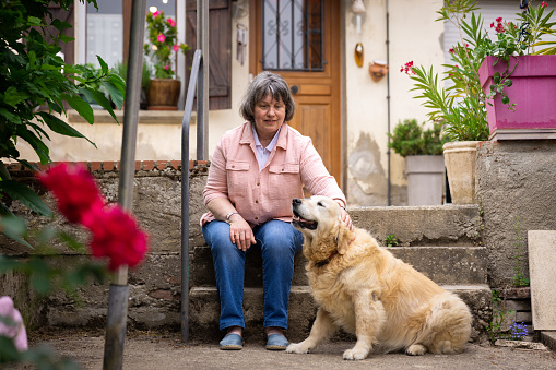 A mature woman who is blind, sitting on steps in the back garden of her home in Castelferrus, France. She is stroking her guide dog and smiling.