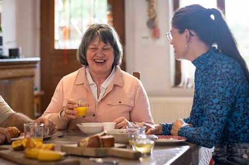 A woman and her daughter enjoying breakfast in their home kitchen together in Castelferrus, France. They are sitting around a dining table and laughing while enjoying a fresh fruit juice. The mother of the family is blind.