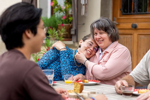 A family and the daughter's boyfriend enjoying a homemade sponge cake and drink together outdoors at an outdoor dining table in Castelferrus, France. They are all sitting around the table and the mother who is blind is embracing her daughter and smiling while they all talk with each other.