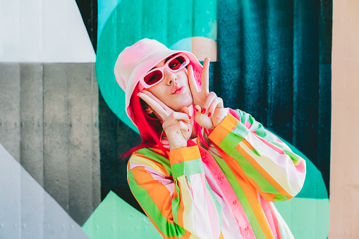 Vanilla Girl. Kawaii vibes. Candy colors design. Young woman with pink hair and sunglasses in Bucket hat and multicolor strippled shirt making V sign by fingers. Anime Urban street fashion