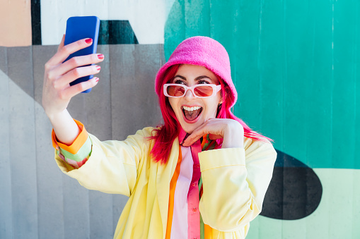 Excited pink hair woman screaming while taking a selfie photo outdoors. Emotional hipster fashion woman in bright clothes, pearl pink sunglasses, bucket hat taking selfie photo on the phone camera.