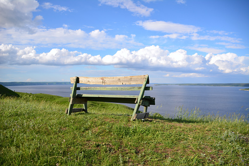 A bench on a top of grassy hill overlooking a body of water with cloudscape