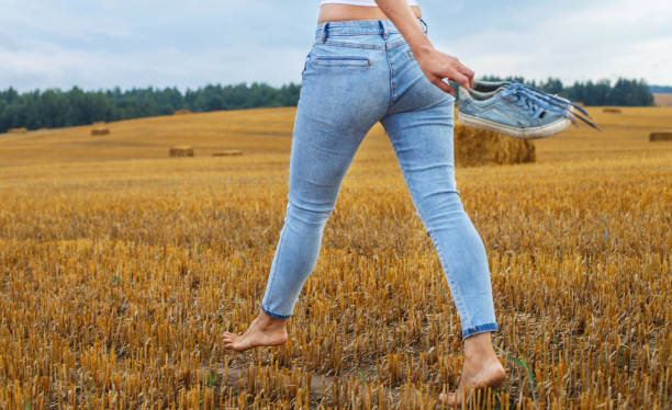 barfüßiges mädchen mit turnschuhen in der hand, das mit heuhaufen und ballen auf dem landwirtschaftlichen feld läuft - child human foot barefoot jeans stock-fotos und bilder