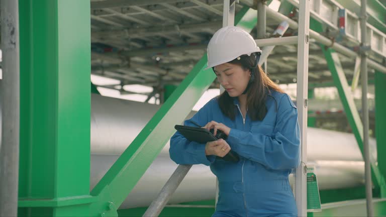 Front view: Beautiful pregnant mid-adult Asian woman management engineer in a blue safety uniform standing at big repaired pipes for inspection and recording in a digital tablet while caring for her baby in the stomach