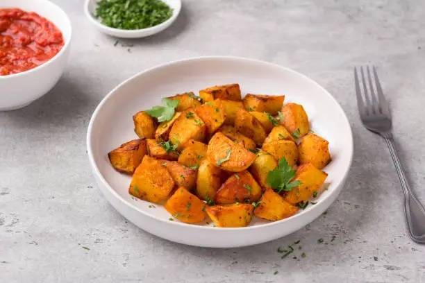 Photo of Traditional Spanish potato, patatas bravas with smoked paprika, spicy tomato sauce and parsley in a white bowl on a gray stone background, top view. Delicious homemade food