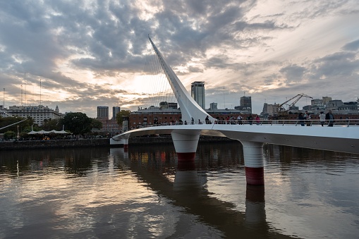 Buenos Aires, Argentina - April 12, 2023: The Women's Bridge or Puente De La Mujer, a counterweight pylon pedestrian cable-stayed bridge in Puerto Madero with Sunset Sky and Urban Highrise Buildings in the Background