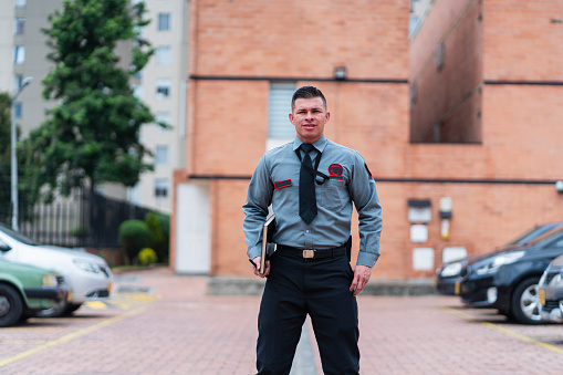 portrait of average age Latino man in his 30s dressed in security guard uniform is inside the building he works for taking bills for vehicles parked in the parking lot