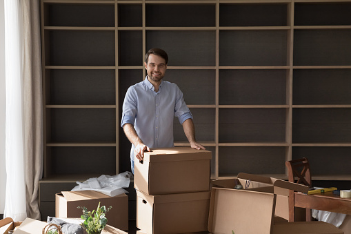 Cheerful satisfied property buyer man posing at stack of cardboard relocation boxes in new apartment, looking at camera with happy smile, packing stuff, unpacking containers, Home owner portrait