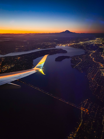 A breathtaking view of Seattle Washington from above just before the sun rises on a crystal clear day. Just off the horizon Mt. Ranier shoots into the orange and blue sky. While the foreground is dotted with the lights of the city and to gorgeous waterways of the area. A wing from the plane enters the top left of the frame.
