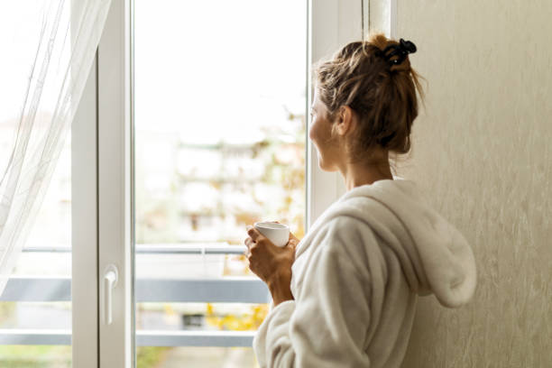 istock Woman at home drinking coffee wearing robe looking through window 1485494860