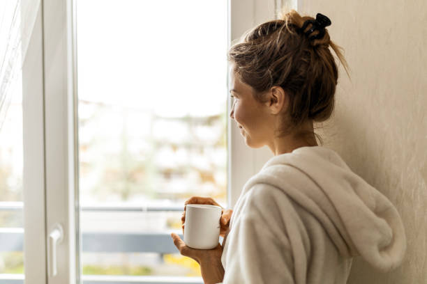 istock Young woman at home drinking coffee wearing robe looking through window 1485494910