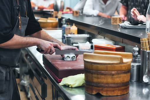 Close-up of a chef's hands with a knife slicing through an uramaki sushi roll with rice and seaweed at a sushi bar with people in the background at the counter