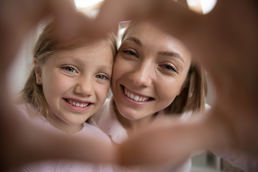 Joyful happy mom and pretty little daughter kid with toothy smile making hand heart frame, joining fingers, showing symbol of love, gratitude, family, kindness, looking at camera. Close up portrait