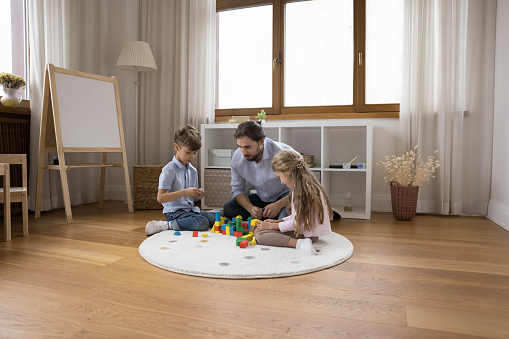 Young caring loving dad entertaining two little sibling kids, constructing toy tower, castle from heap of building blocks on warm floor in home playroom. Male sitter, daycare teacher watching children