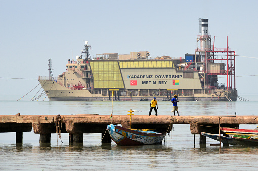 Bissau, Guinea-Bissau: moored in the harbor, the Turkish power-ship Metin Bey (IMO: 9034779) of the Karmarine Karadeniz Denizcilik company produces electrical power for the capital, a pylon at the stern is the origin of power cables that reach the shore. In the foreground fishermen and traditional fishing boats on the pier. Powerships are a solution for countries where blackouts and power outages are common. Such vessels can operate blissfully unaffected by the governance, security and other issues that often render conventional shore-based powers stations inoperational.