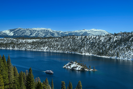 Winter view of Emerald Bay with Fannette Island in the middle with passing paddleboat and snow covered mountains in background.\n\nTaken from the Western Shore of Lake Tahoe, California, USA  looking East.